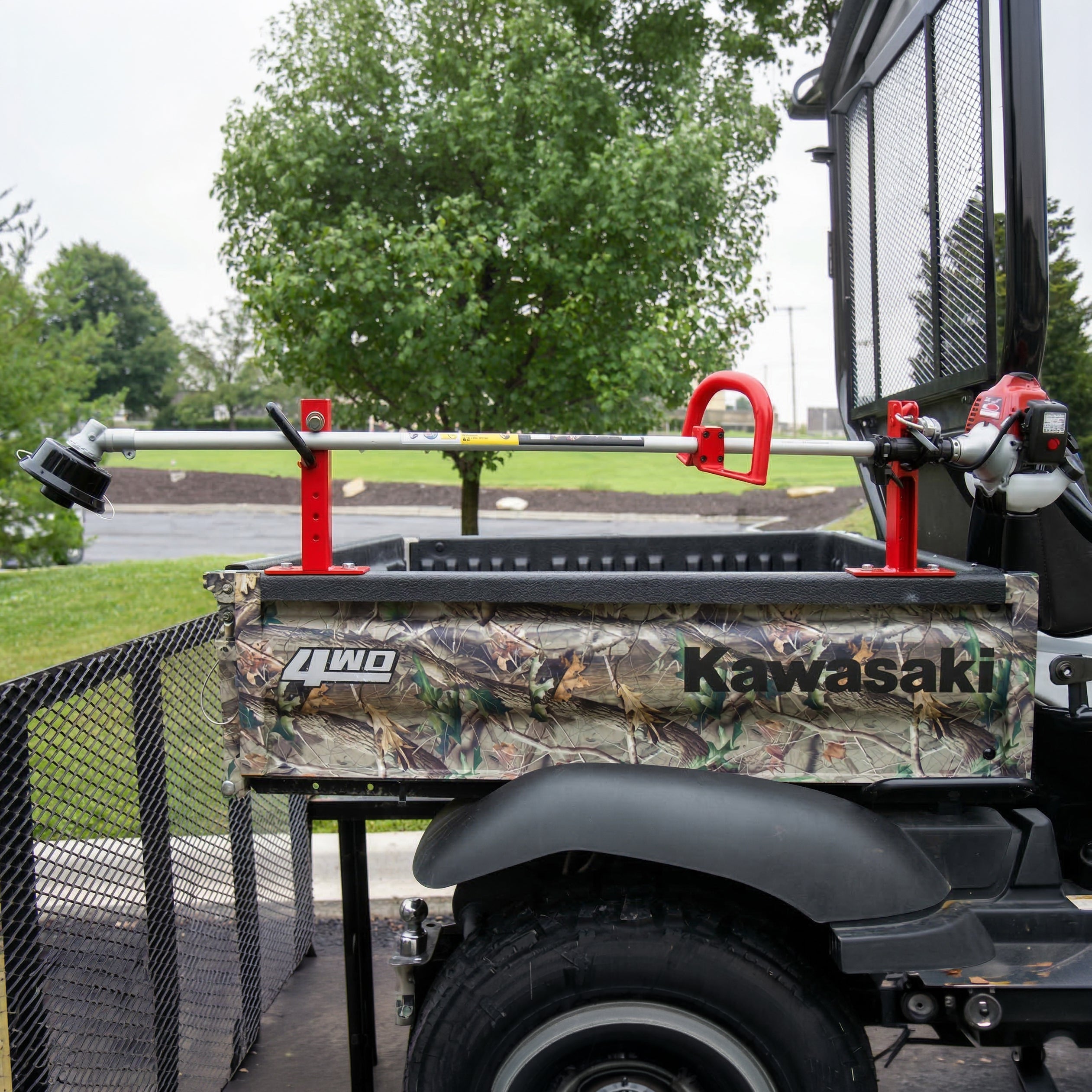 A professional-grade 1TR trimmer rack securely mounted to the side rail of a camouflage Kawasaki UTV. The rack holds a red gas-powered string trimmer horizontally, with the engine positioned to the right and the trimmer head to the left, demonstrating a secure and organized equipment storage solution for landscaping and utility work.