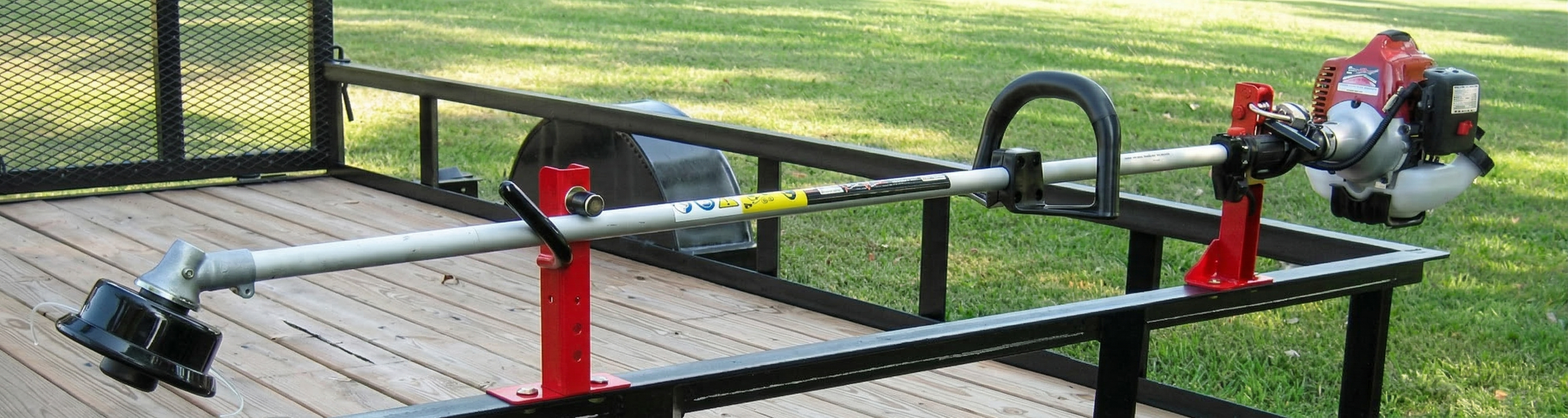 A Stihl string trimmer securely held by a Jungle Jim’s 1TR rack on an open utility trailer with professional striped turf background.